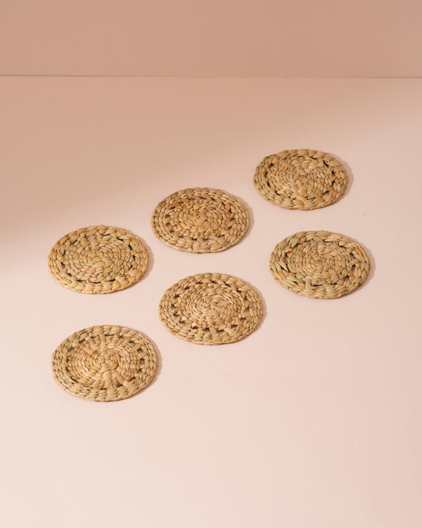Six beige handwoven Kouna grass coasters on pink surface